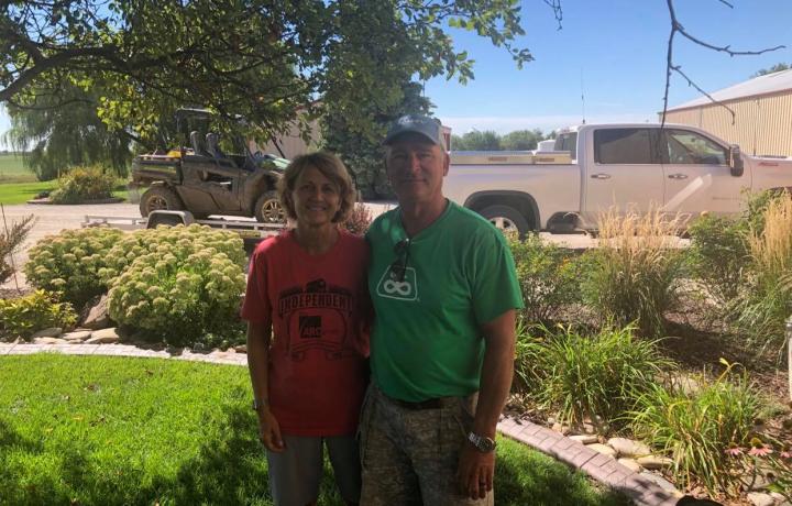 Ron and Rona Volkmer are pictured in their yard, posing in the shade of a large tree surrounded by landscaping. Both wear colorful T-shirts and khaki shorts.