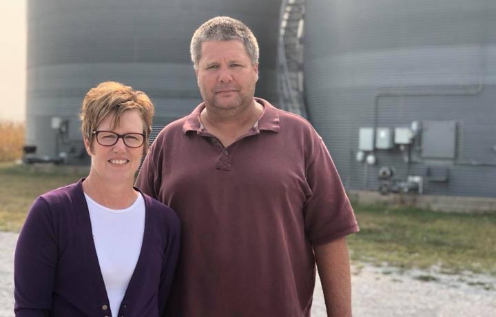 Brian and Nicole Nedrow are pictured near bins on their farm near Geneva. It is a bright September day. Nicole wears a purple cardigan and white T-shirt. Brian wears a purple polo shirt.