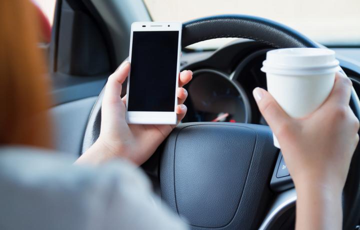 This picture is a close-up shot of a steering wheel in a car. In the female driver's hands are a cell phone and a cup of coffee, held as she drives.