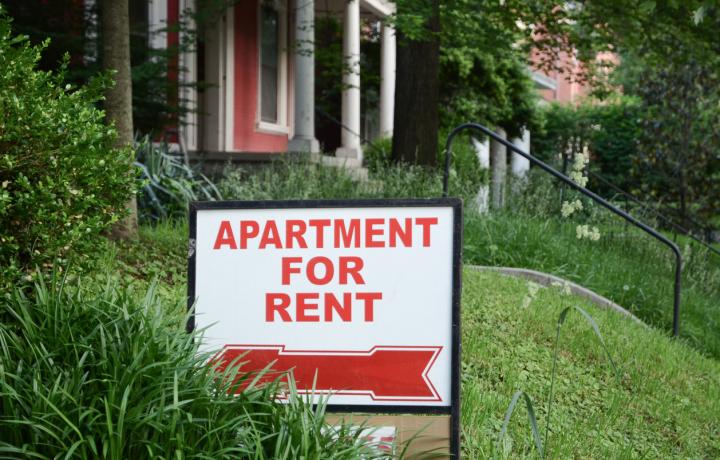 A sign staked into a grassy slope advertises 'apartment for rent.' A large home with a broad front porch is pictured in the background.