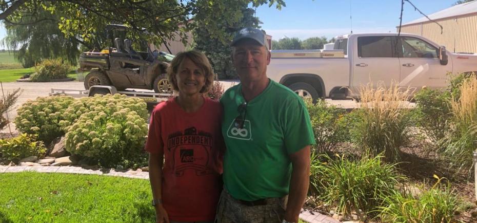 Ron and Rona Volkmer are pictured in their yard, posing in the shade of a large tree surrounded by landscaping. Both wear colorful T-shirts and khaki shorts.