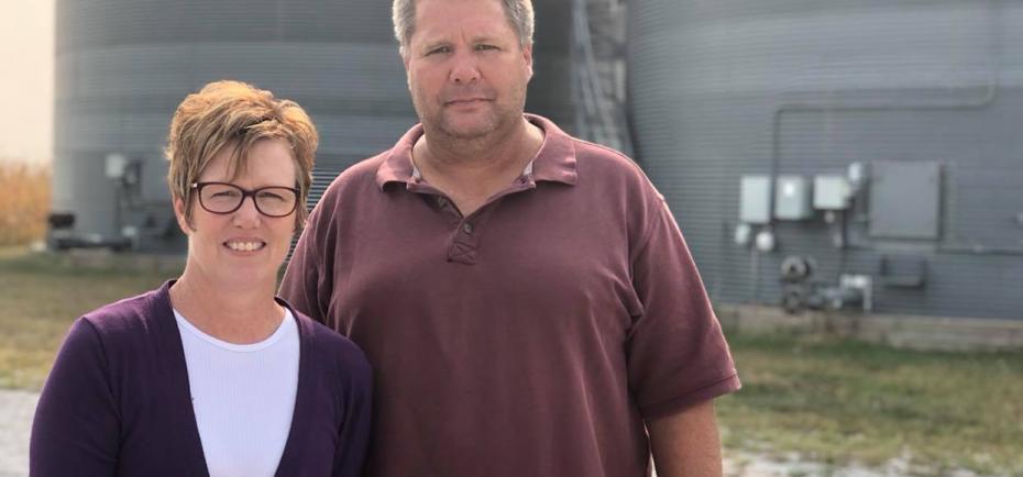Brian and Nicole Nedrow are pictured near bins on their farm near Geneva. It is a bright September day. Nicole wears a purple cardigan and white T-shirt. Brian wears a purple polo shirt.