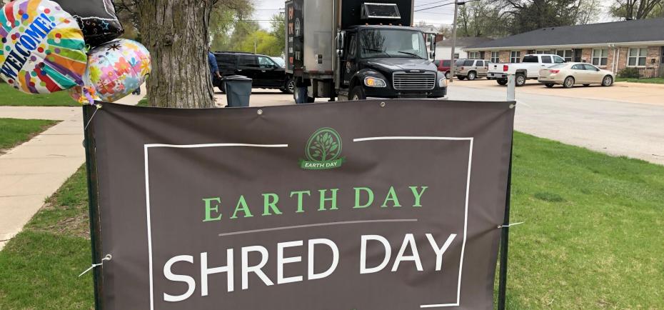 A gray and green Shred Day for Earth Day banner is shown in the foreground. The Paper Tiger mobile shredding truck sits in the background.