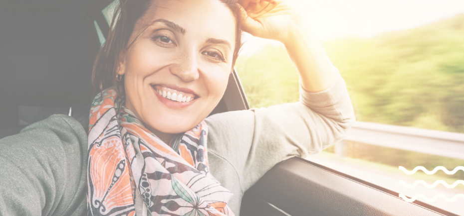 A woman in a gray sweater and colorful scarf smiles behind the wheel of a car on a sunny day.