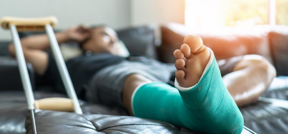 A young man with a broken foot reclines on a sofa wearing a green foot cast. A single crutch is pictured in the foreground.