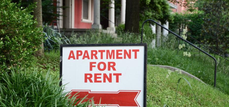 A sign staked into a grassy slope advertises 'apartment for rent.' A large home with a broad front porch is pictured in the background.
