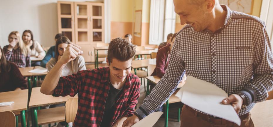 A teacher hands a young man his school paper, which earned a good grade. The young man, with dark hair and a plaid shirt, makes a jubilant expression as he receives it on his desk.