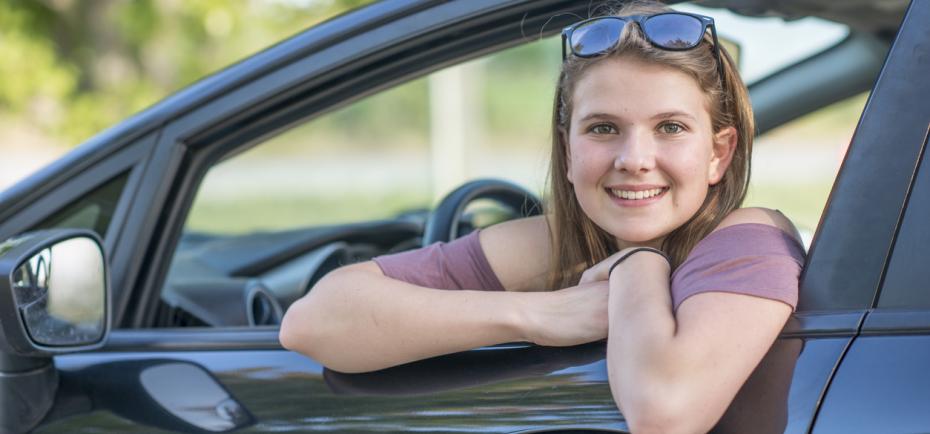 A teenage girl smiles out of an open window of the car she is about to drive. She is wearing a short-sleeved mauve T-shirt and has sunglasses on top of her head.
