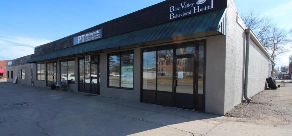 A one-story retail/commercial building is pictured. It's for sale and sits along G St. and Highway 41 in Geneva. It has a gray and black color scheme. 