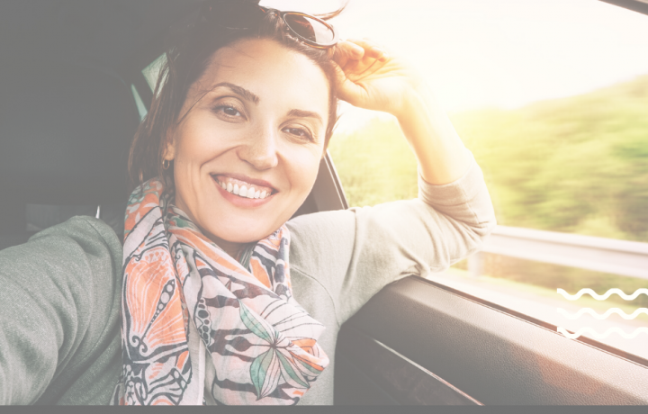 A woman in a gray sweater and colorful scarf smiles behind the wheel of a car on a sunny day.