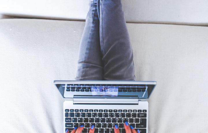 A young woman works on a laptop computer while lying comfortably on top of a bed. Her bright green sweater and blue jeans pop against the white comforter.