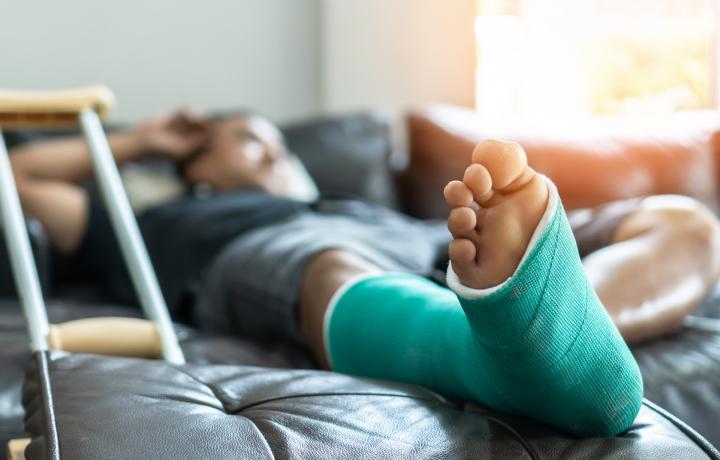 A young man with a broken foot reclines on a sofa wearing a green foot cast. A single crutch is pictured in the foreground.