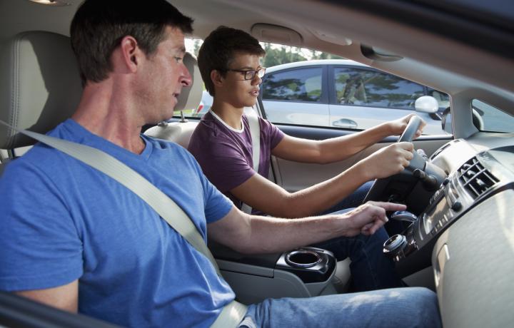 A father in the passenger seat leans over to point out a driving tip to his young son in the driver seat of the car.