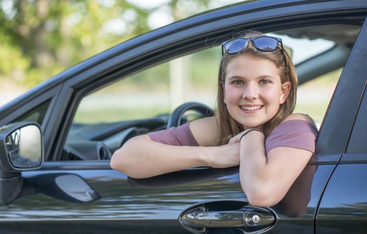 A teenage girl smiles out of an open window of the car she is about to drive. She is wearing a short-sleeved mauve T-shirt and has sunglasses on top of her head.