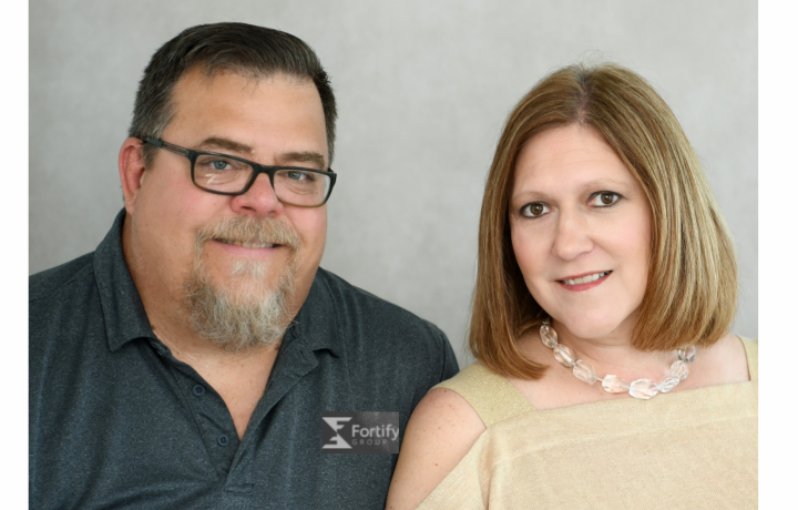New Fortify Group team members Jim and Kate Manes are pictured in this professional portrait. Jim wears glasses and a gray polo; Kate has on a pale cream sweater.