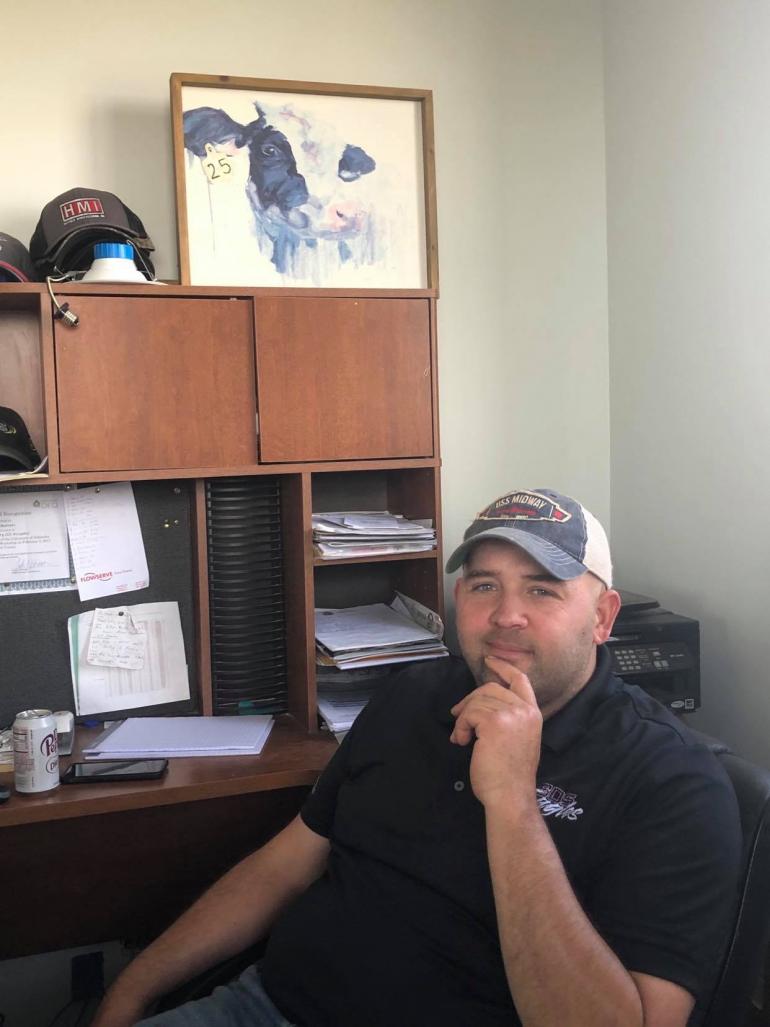 Brett Beavers is pictured in the office of Beavers Dairy near Shickley. He wears a black polo shirt and ball cap; a picture of a Holstein cow is placed on the cabinet nearby.