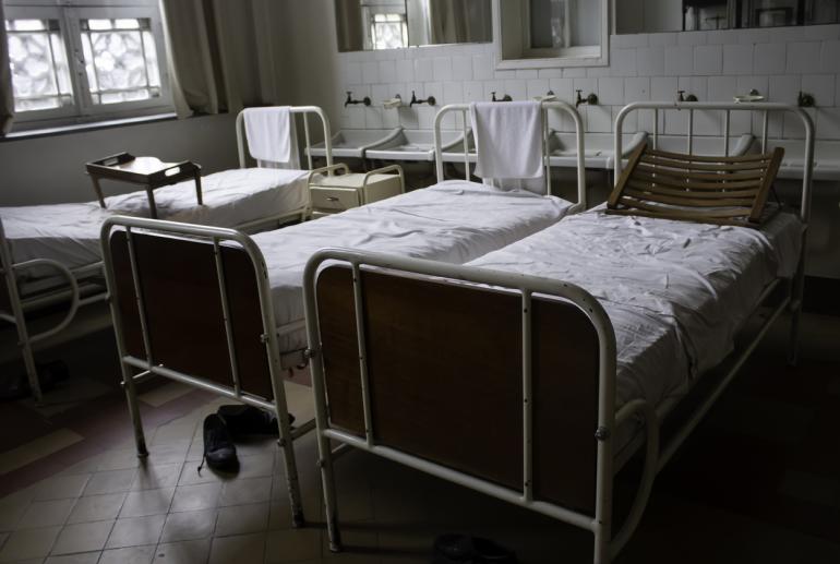 1950s hospital beds are shown in an unnamed ward. They are made of white iron with white sheets.