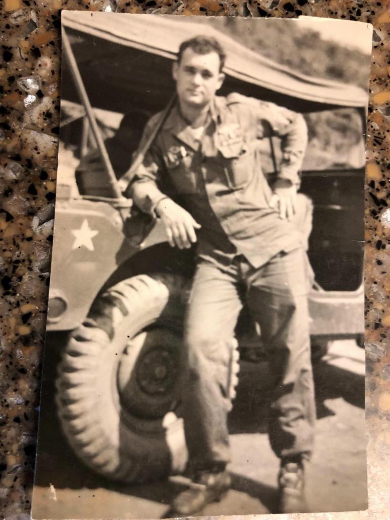 young Tanny Reinsch is picture in the Korean War. In his soldier's uniform, he leans against an army jeep for the picture.