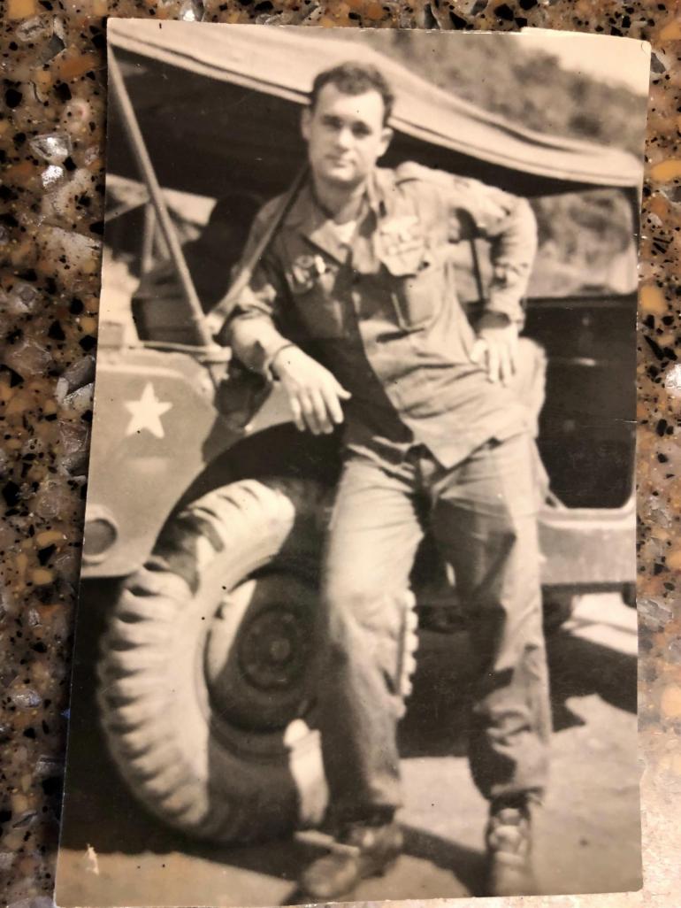  young Tanny Reinsch is picture in the Korean War. In his soldier's uniform, he leans against an army jeep for the picture.