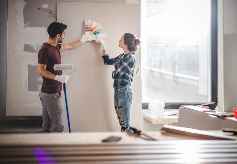 A young man and women prepare to paint a living space wall. She fans a handful of paint chips; he holds a paint roller with a long handle.