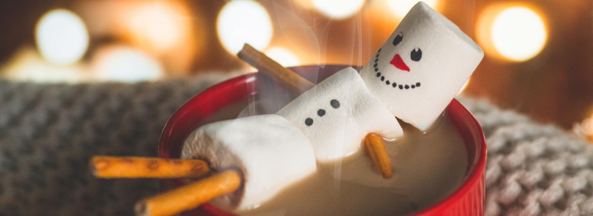 In this cozy winter photo, a stick figure made from marshmallows and cinnamon sticks appears to be relaxing in a pool (cup) of hot chocolate. A bright red mug and white lights in the background complete a holiday feel.