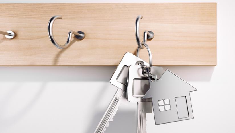 A ring of shiny silver keys hang on a key hook on a maple board. The keys include one key in the shape of a house.